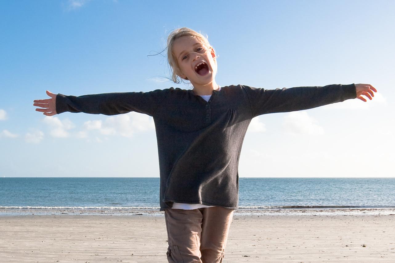 Mädchen steht am Strand mit ausgebreiteten Armen, lächelt und blickt in die Kamera gegen blauen Himmel und Meer.