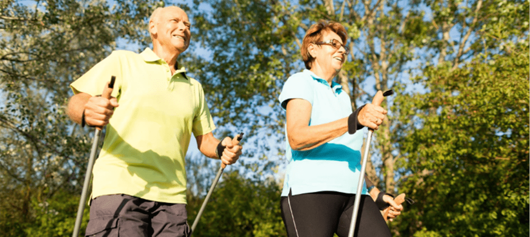 Paar beim Nordic Walking in der Natur, lächelnd mit Stöcken, umgeben von Bäumen und Sonnenschein.