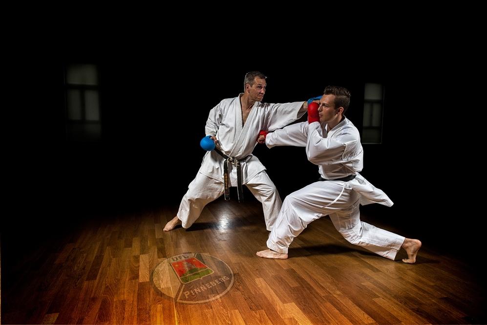 Zwei Karatekämpfer in weißen Anzügen üben Techniken auf einem Holzboden in einem dunklen Raum.